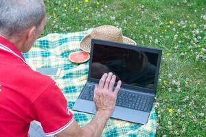 midden- oud Mens met mobiel telefoon en laptop leugens Aan een deken in groen gras foto