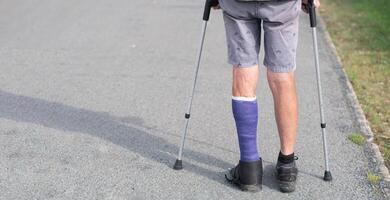 Mens met een gebroken been wandelingen naar beneden de straat, zijn links been is in een vorm, de Mens beweegt met de helpen van krukken foto