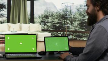 ondernemer werken met groene scherm Aan laptop en tablet, zittend Bij bureau in zijn chique modern alpine cabine. rijk zakenman onderzoeken geïsoleerd mockup wordt weergegeven Aan twee gadgets, miljardair. foto