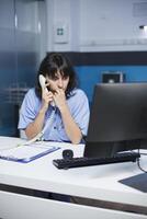 een moe Kaukasisch vrouw dokter in een modern ziekenhuis kantoor, werken Aan een computer, Holding een telefoon, en omringd door medisch apparatuur. een verpleegster in blauw scrubs sprekend Aan een vaste telefoon. foto