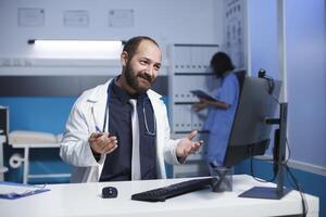 gebaard Kaukasisch Mens vervelend een laboratorium jas in een kantoor verloofd in een video telefoontje Aan een bureaublad pc, bespreken medisch zaken. andere gezondheidszorg professionals werk in de achtergrond. foto