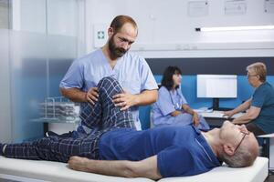 gepensioneerd oud Mens met knie letsel ontvangen osteopathisch bijstand voor fysiek herstel. gezondheidszorg assistent masseren senior geduldig met been spier pijn, helpen naar verlichten druk. foto