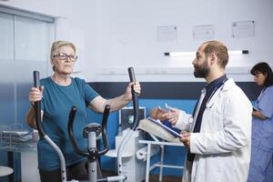 mannetje dokter pratend naar senior geduldig aan het doen fysiek werkzaamheid naar herstellen van letsel met elektrisch fiets. senior vrouw gebruik makend van stationair fiets voor welzijn en herstel Bij fysiotherapie kliniek. foto