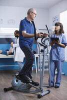 ouderen mannetje geduldig ondergaan fysiotherapie Aan stationair fiets, met bijstand van verpleegster in blauw schrobt. medisch assistent gebruik makend van elektronisch tablet assists senior inwoner met fysiek therapie. foto