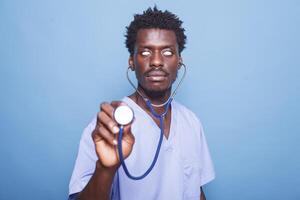 in studio, speels mannetje verpleegster is geklemd een stethoscoop en rollend zijn ogen. tegen blauw achtergrond, Afrikaanse Amerikaans medisch assistent houdt een cardiologie instrument voor een hartslag inspectie. foto