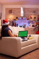 vrouw persoon resting Aan sofa in Kerstmis versierd keuken Holding groen scherm bespotten omhoog chroma sleutel laptop computer met geïsoleerd Scherm. volwassen vrouw genieten van Kerstmis vakantie vieren winter seizoen foto
