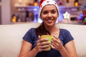 portret van glimlachen vrouw Holding kop van koffie in handen gedurende kersttijd zittend Aan bankstel in Kerstmis versierd keuken. volwassen persoon genieten van winter seizoen vieren Kerstmis vakantie foto