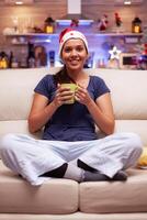 portret van glimlachen vrouw vervelend rood de kerstman hoed zittend in lotus positie Aan sofa in Kerstmis versierd keuken op zoek in camera genieten van Kerstmis vakantie. vrouw vieren winter seizoen foto