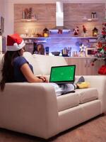 meisje op zoek Bij bespotten omhoog groen scherm chroma sleutel laptop met geïsoleerd Scherm aan het liegen Aan sofa in Kerstmis versierd keuken. vrouw genieten van winter seizoen vieren Kerstmis vakantie foto