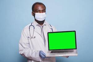 portret van gezondheidszorg specialist draag- draadloze computer weergeven blanco Chroma sleutel mockup sjabloon. Afrikaanse Amerikaans mannetje arts grijpen digitaal laptop met groen scherm. foto