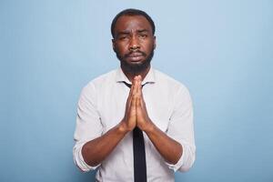 staand over- een blauw achtergrond, een Afrikaanse Amerikaans zakenman bidt met zijn handen samen in hoop. verdrietig medewerker, op zoek heel emotioneel, maakt een hand- gebaar voor vergiffenis. foto
