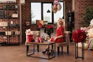 vrouw zittend in leven kamer gevulde met romantisch presenteert, ontvangen Valentijn s dag cadeaus van vriendje gedurende liefde vakantie. schattig blond vrouw in rood elegant jurk genieten van februari 14 verrassingen foto