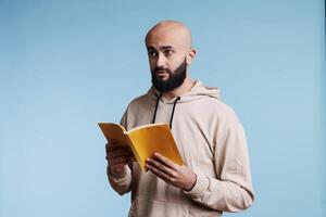 jong Arabisch Mens Holding zachte kaft boek en overweegt verhaal terwijl op zoek weg. Arabisch persoon lezing roman paperback met geel Hoes terwijl staand met attent uitdrukking foto