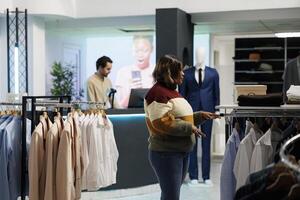 Afrikaanse Amerikaans vrouw selecteren kleding in elegant kleding boetiek, zoeken overhemd grootte Aan rek. kleding op te slaan klant controle formeel slijtage Aan hangers terwijl boodschappen doen in stopcontact foto