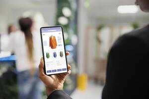 cliënt gebruik makend van kleding online web op te slaan toepassing Aan smartphone, herzien mode items en buying producten. Afrikaanse Amerikaans vrouw op zoek Bij modieus kleren lijn Aan digitaal winkel. foto