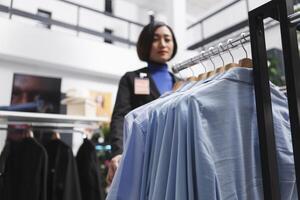 blauw gewoontjes overhemden hangende Aan kleding rek terwijl Aziatisch vrouw verkoper staand in winkelcentrum kleding op te slaan selectief focus. elegant kledingstuk Aan hangers in afdeling centrum winkel detailopname foto