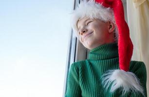 kinderen dromen concept. vriendschap. een eenzaam weinig jongen in een de kerstman claus hoed is verdrietig Bij de venster aan het wachten voor geschenken, op zoek uit de venster, de kind is aan het wachten voor de kerstman claus. foto