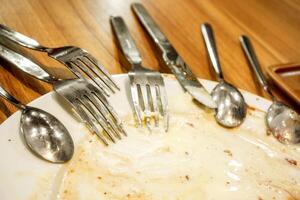 roestvrij lepel, mes en vork zetten Aan wit keramisch vuil schotel van aan het eten voedsel en houten tafel. foto