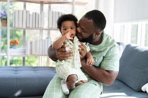 gelukkig Afrikaanse Amerikaans Mens Speel met weinig zuigeling kleuter kind in leven kamer bank. ouder en weinig kind ontspannende Bij huis. ouders en kleuter kind meisje genieten van uitgeven tijd Bij huis. kinderopvang foto