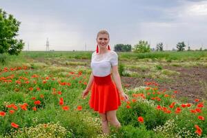blond jong vrouw in rood rok en wit shirt, rood oorbellen is in de midden- van een papaver veld. foto