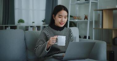 portret van gelukkig jong Aziatisch vrouw zitten Aan sofa drinken koffie of thee terwijl werken creatief baan van huis Aan laptop met online netwerk, nemen pauze, glimlachend foto