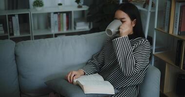 portret van gelukkig jong Aziatisch vrouw drinken ochtend- koffie of thee en lezing in leven kamer Bij huis Aan weekend. vrije tijd en levensstijl, gratis tijd foto