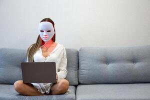 een mooi meisje met een LED masker Aan haar hoofd werken Bij huis Aan een laptop. huis huid zorg concept. foto