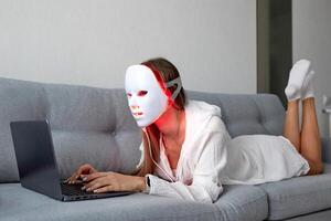 een mooi meisje met een LED masker Aan haar hoofd werken Bij huis Aan een laptop. huis huid zorg concept. foto