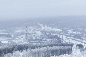 ijzig winter landschap - een ver weg metallurgisch fabriek in een vallei in de midden- van besneeuwd bossen in een ijzig nevel foto