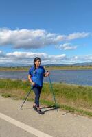 buitenshuis liefhebber met wandelen polen naast een rustig water lichaam Aan een helder dag, spaans latina vrouw wandelen met trekking polen in de ebro delta natuurlijk park, tarragona, Catalonië, Spanje foto