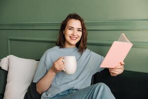 gelukkig intelligent jong middelbare leeftijd vrouw lezing een boek, genieten van ontspanning en een kop van thee in haar Reserve tijd foto