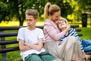 gelukkig moeder is zittend met haar zonen Aan bank in park. een jongen is beledigd en pruilend foto