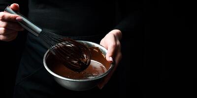 een kom van gesmolten chocola en een keuken vliegenmepper zijn gehouden door handen. keuken gebruiksvoorwerpen. Aan een zwart achtergrond. foto