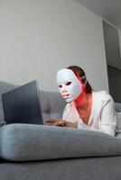 een mooi meisje met een LED masker Aan haar hoofd werken Bij huis Aan een laptop. huis huid zorg concept. foto