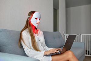 een mooi meisje met een LED masker Aan haar hoofd werken Bij huis Aan een laptop. huis huid zorg concept. foto
