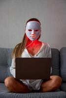 een mooi meisje met een LED masker Aan haar hoofd werken Bij huis Aan een laptop. huis huid zorg concept. foto