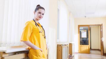 blij jong verpleegster. glimlachen jong meisje in geel uniform, en op zoek Bij camera, in voorkant van venster kliniek. de concept van Gezondheid foto