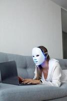 een mooi meisje met een LED masker Aan haar hoofd werken Bij huis Aan een laptop. huis huid zorg concept. foto