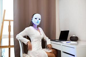 een mooi meisje met een LED masker Aan haar hoofd werken Bij een laptop. huis huid zorg concept. foto