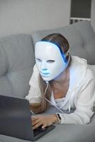 een mooi meisje met een LED masker Aan haar hoofd werken Bij huis Aan een laptop. huis huid zorg concept. foto