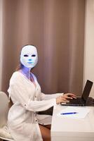 een mooi meisje met een LED masker Aan haar hoofd werken Bij een laptop. huis huid zorg concept. foto