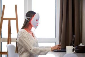 een mooi meisje met een LED masker Aan haar hoofd werken Bij een laptop. huis huid zorg concept. foto