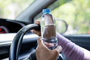 Aziatische vrouw die een fles vasthoudt om water te drinken tijdens het autorijden. plastic warmwaterkruik brand veroorzaken. foto