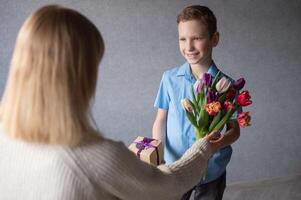 schattig jongen glimlachen en op zoek Bij mama, geven bloemen en cadeaus voor moeder dag foto