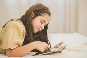 kind houdende Aan bed gebruik makend van tablet voor zelf aan het leren onderwijs Bij huis. schattig meisje Speel vakantie spellen Aan touch screen apparaat. foto