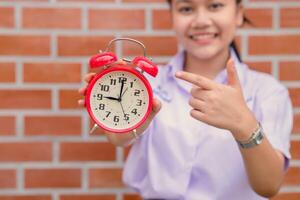 gelukkig Aziatisch school- tiener meisje glimlachen hand- richten Bij alarm klok voor keer naar wakker naar onderwijs prioriteit.oproep voor aan het leren concept foto