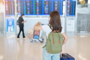 jong vrouw met zak en bagage controle vlucht tijd informatie bord in Internationale luchthaven, voordat controleren in. reis, vakantie, reis en vervoer concept foto