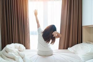 gelukkig vrouw uitrekken Aan bed na wakker worden omhoog, jong volwassen vrouw stijgende lijn armen en op zoek naar venster in de vroeg ochtend. vers kom tot rust en hebben een mooi hoor dag concepten foto