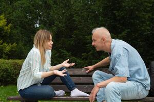 Mens met grijs haar- en Kaukasisch vrouw zijn zittend Aan een bank in de park tegenover en pratend foto