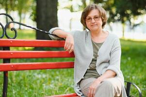 elegant ouderen vrouw in de overhemd is zittend Aan de bank in een park Aan een warm dag foto